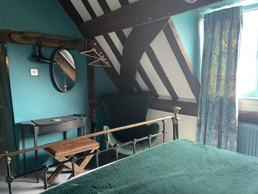 Una habitación con una cama, un sillón tapizado en verde, una mesita, un espejo y una ventana con cortinas estampadas.