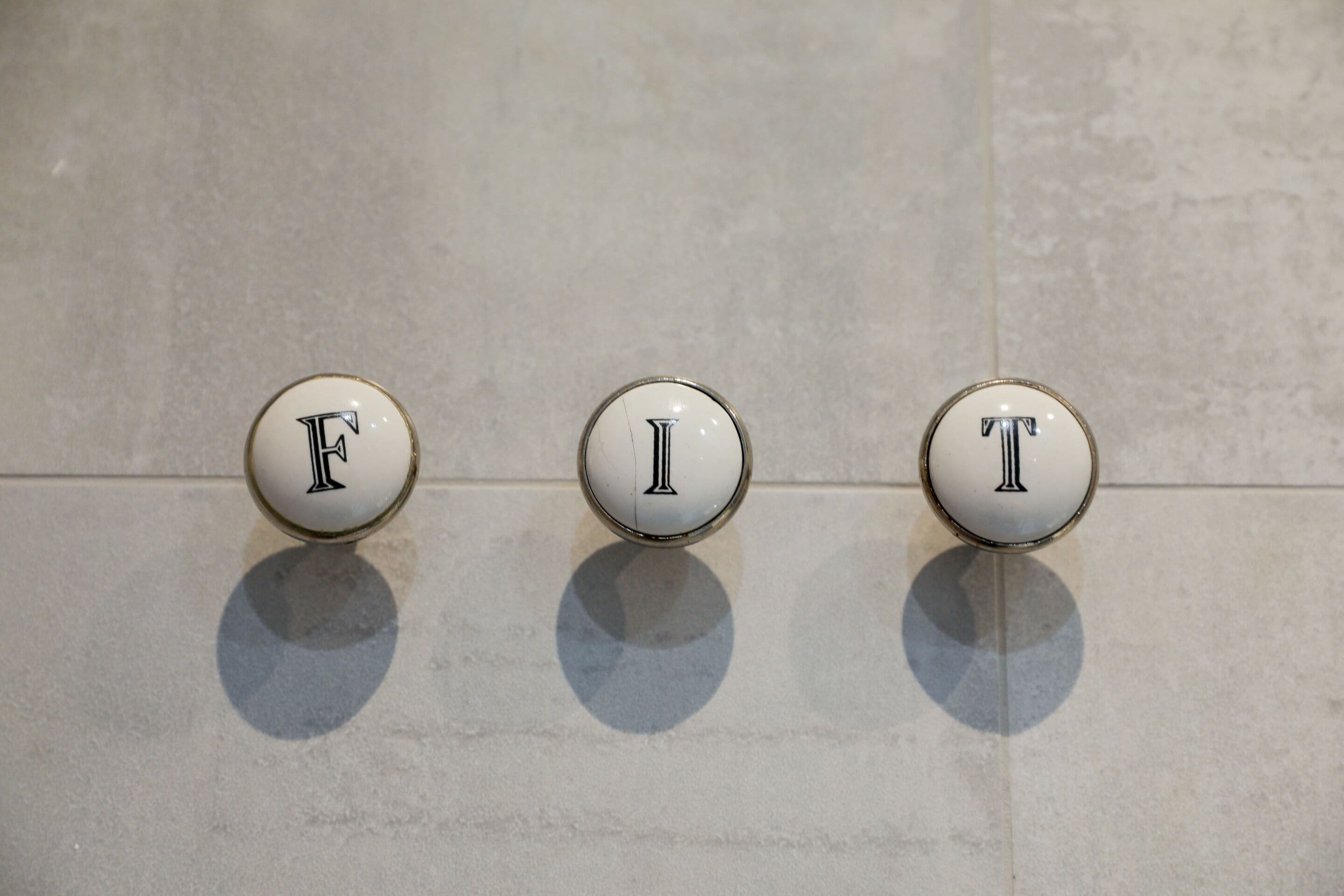 Trois boutons ronds et blancs, portant les lettres « F », « I » et « T », sont fixés sur un mur recouvert de carreaux.