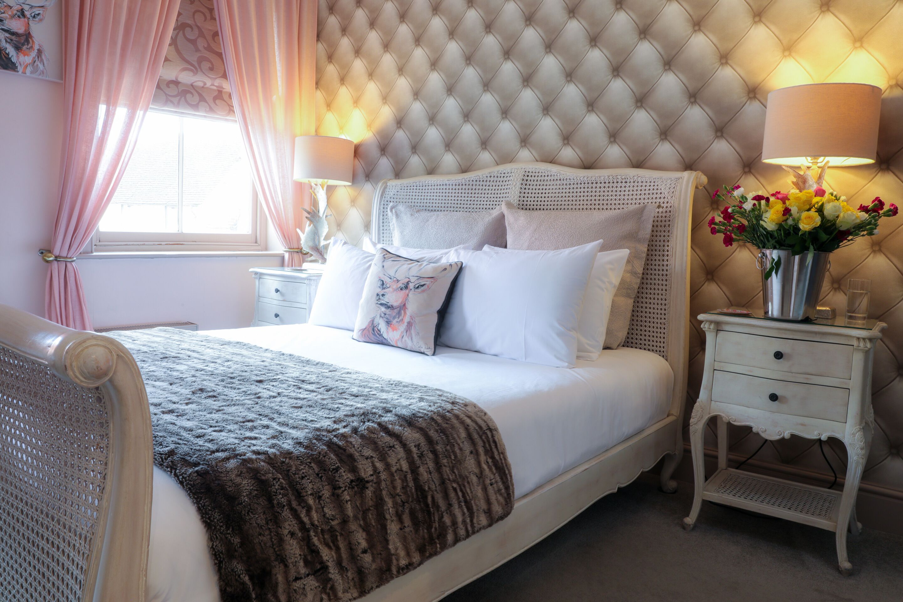 Une chambre à coucher avec une tête de lit capitonnée, du linge de lit blanc, un plaid décoratif, des tables de chevet avec des lampes et un vase rempli de fleurs.