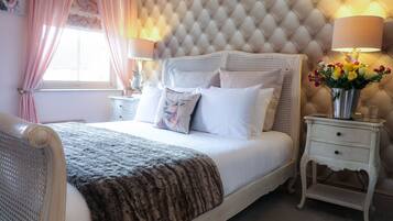 Une chambre à coucher avec une tête de lit capitonnée, du linge de lit blanc, un plaid décoratif, des tables de chevet avec des lampes et un vase rempli de fleurs.