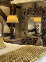 A bedroom with a bed, a nightstand with a lamp, a mirror with an ornate frame, and a decorative sculpture.