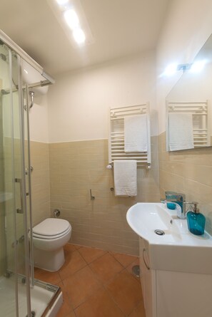 A bathroom with a glass shower enclosure, a white toilet, beige tiled walls, and a white sink with a blue soap dispenser.
