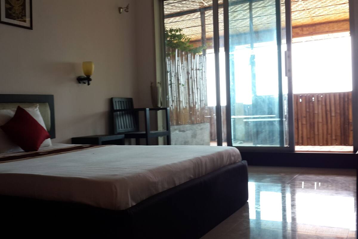 A bedroom with a large bed, a chair, a nightstand, and a window with a view of a bamboo fence.