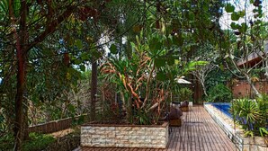 Una terraza de madera con una jardinera de piedra, rodeada de abundante follaje y una pequeña piscina.