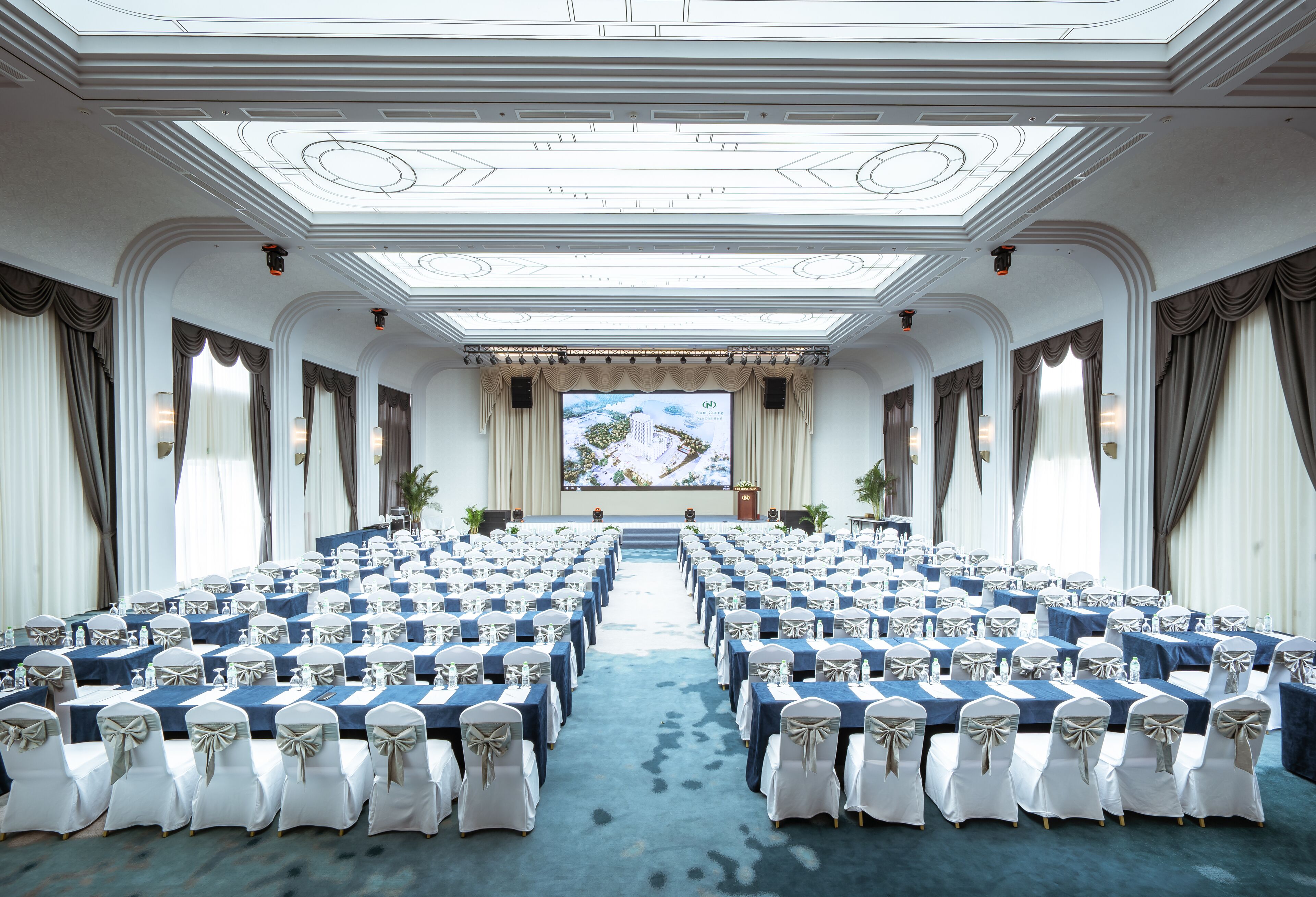Een congreszaal met rijen tafels en stoelen, een groot scherm aan de voorkant en een hoog plafond met decoratieve sierlijsten.