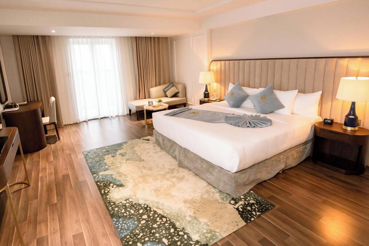 A hotel room with a large bed, two bedside tables with lamps, a TV, a sofa, and a desk with a chair.