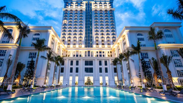 A large hotel with a swimming pool in front, palm trees, and a clear sky. - Nam Cuong Nam Dinh Hotel (Nam Dinh)
