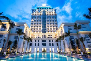 A large hotel with a swimming pool in front, palm trees, and a clear sky.