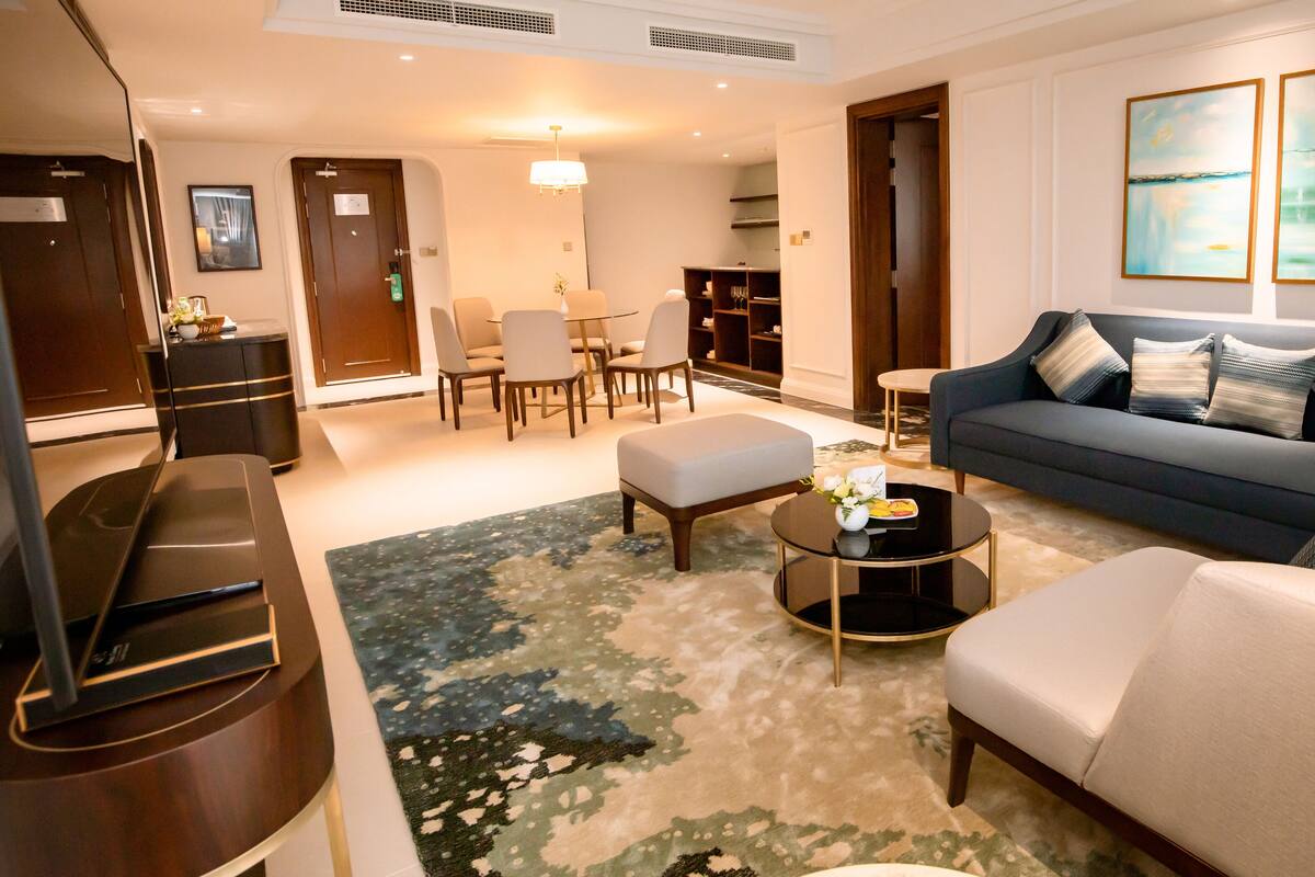 A modern living room with a sofa, armchair, and coffee table. There is a dining area with chairs and a table. The room has a large window and artwork on the wall.