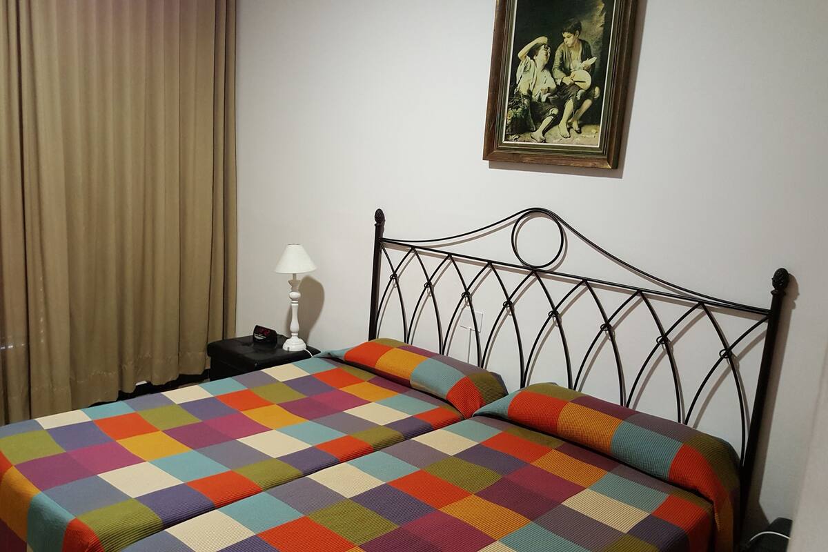 Un dormitori amb llit, cortines, una tauleta de nit, una làmpada i un quadre a la paret.