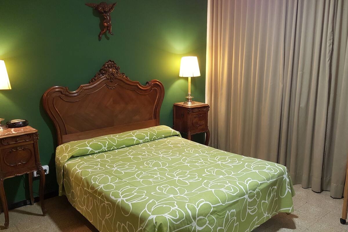 Habitació d'hotel amb paret verda, fusta de capçal de llit, cobrellit floral verd, dues làmpades de nit, taulella de nit de fusta i una cadira de fusta.
