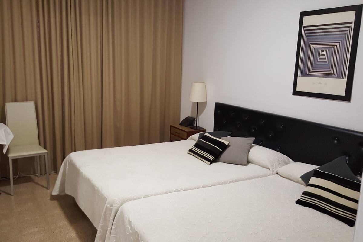 Habitació d'hotel amb dos llits, una taula petita amb una làmpada, una cadira i una obra d'art penjada a la paret.