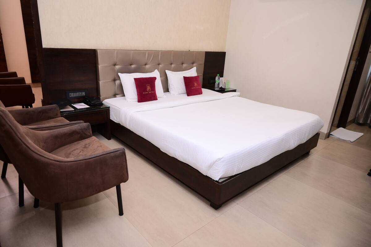 A hotel room with a large bed, two red pillows, a brown armchair, a bedside table with a phone and water bottles, and a nightstand with a lamp.