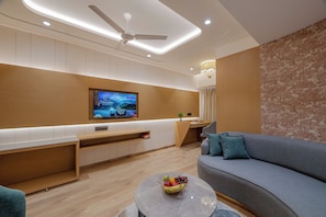 A modern living room with a flat-screen TV mounted on the wall, a sofa, a coffee table with fruit, and a desk area. - Pravasa Hotel Indore (Indore)