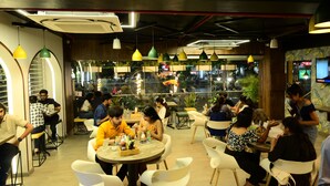 A restaurant with round tables and white chairs, featuring a glass wall with plants and a view of the street.