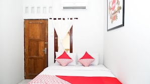 Une chambre avec une porte en bois, des murs blancs, un lit recouvert d’une courtepointe à motifs rouges et blancs, deux oreillers rouges, un rideau blanc et un appareil de climatisation.