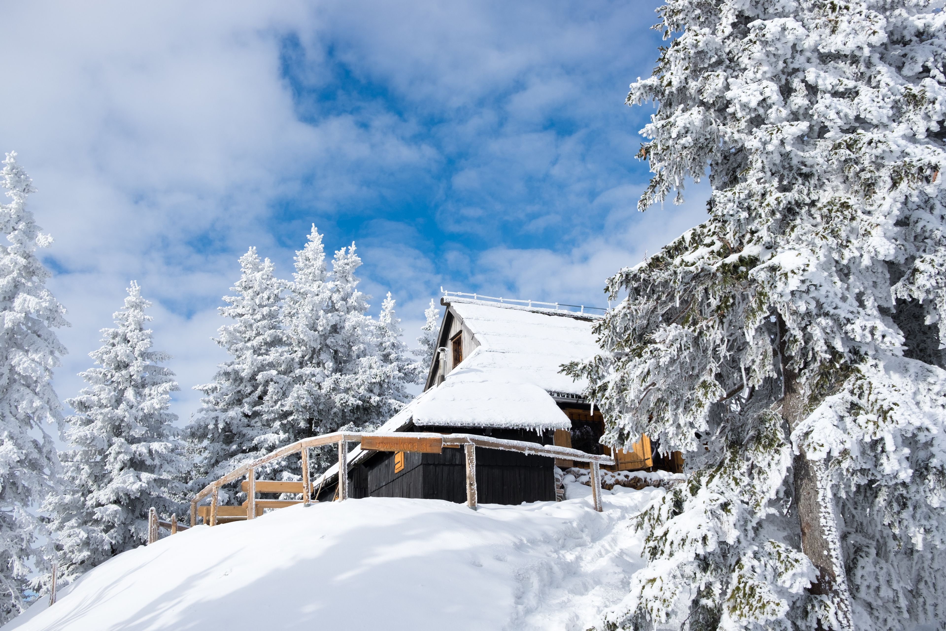 A snow-covered wooden cabin with a wooden deck, surrounded by snow-laden trees, under a partly cloudy sky.