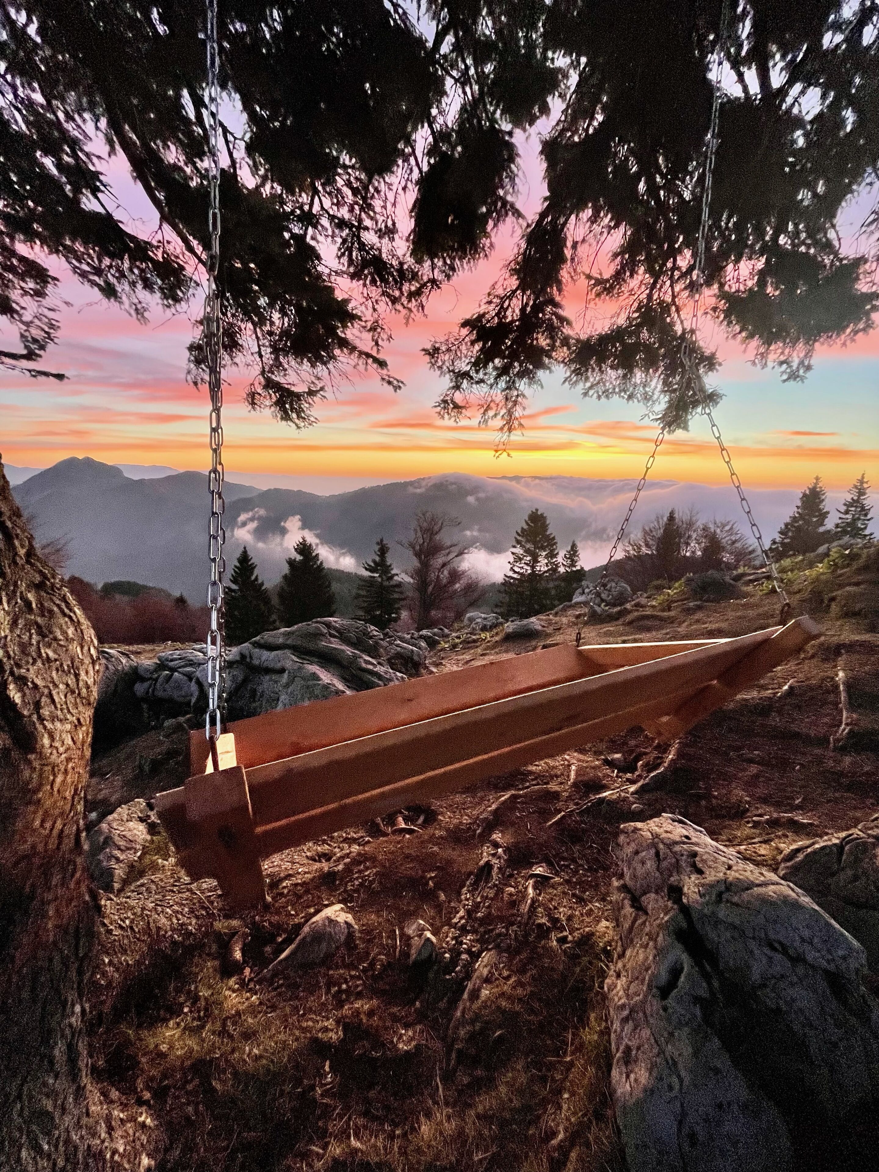 Una panchina di legno su una sporgenza rocciosa con vista sulle montagne e un tramonto sullo sfondo.