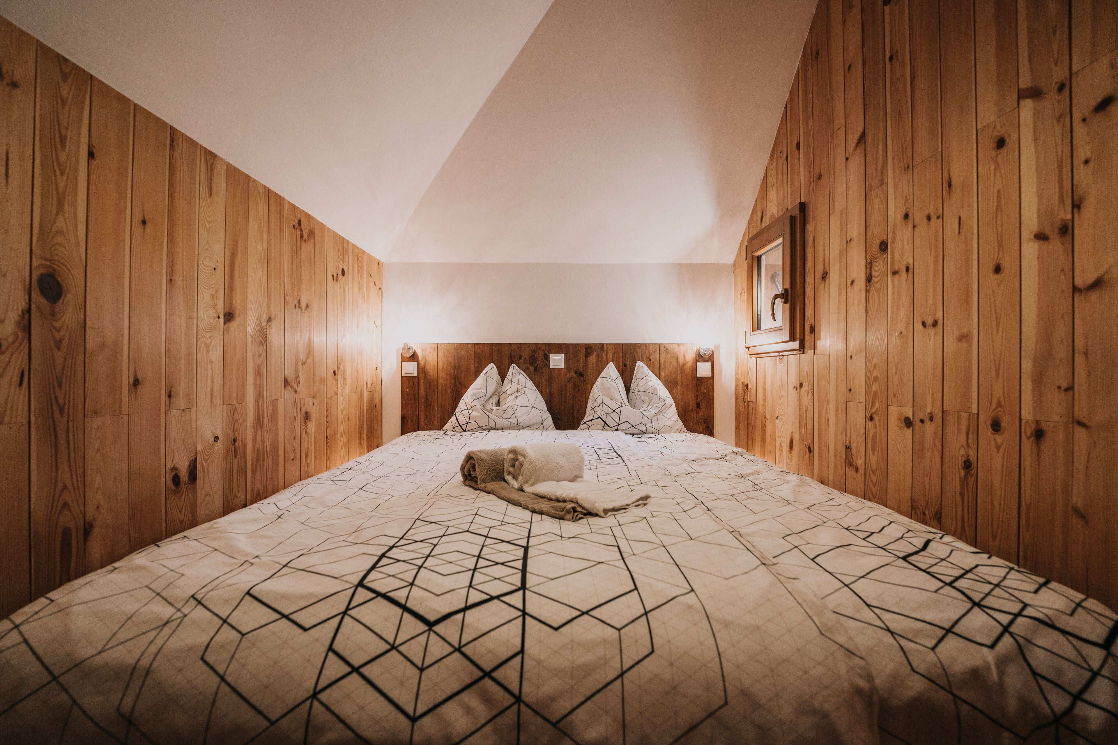 Ein Zimmer mit Holzwänden, einem Bett, einer Bettdecke mit geometrischem Muster und einem gerahmten Bild an der Wand.