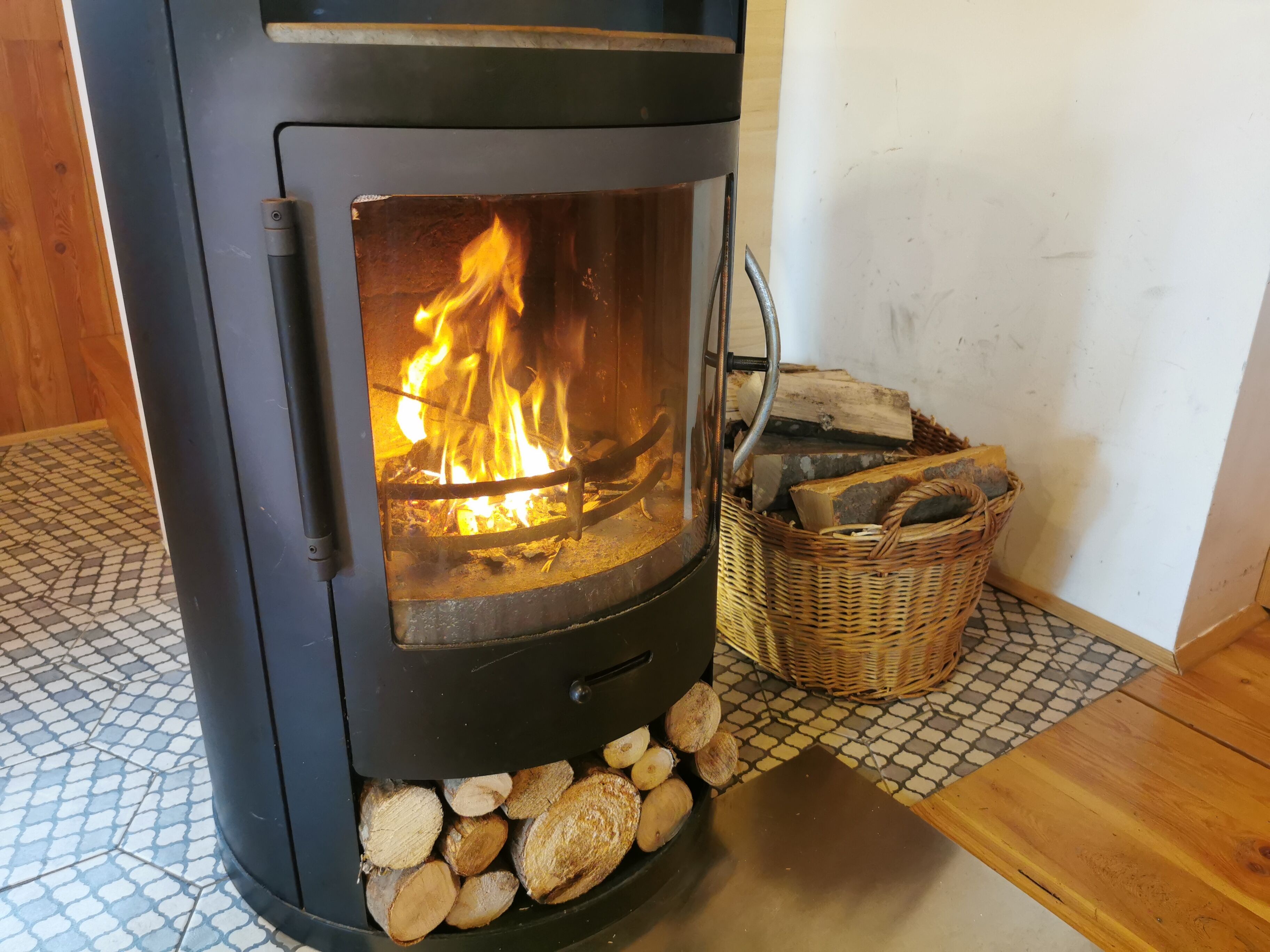 Uma lareira a lenha com fogo crepitando no interior, rodeada por toras e um cesto de lenha.