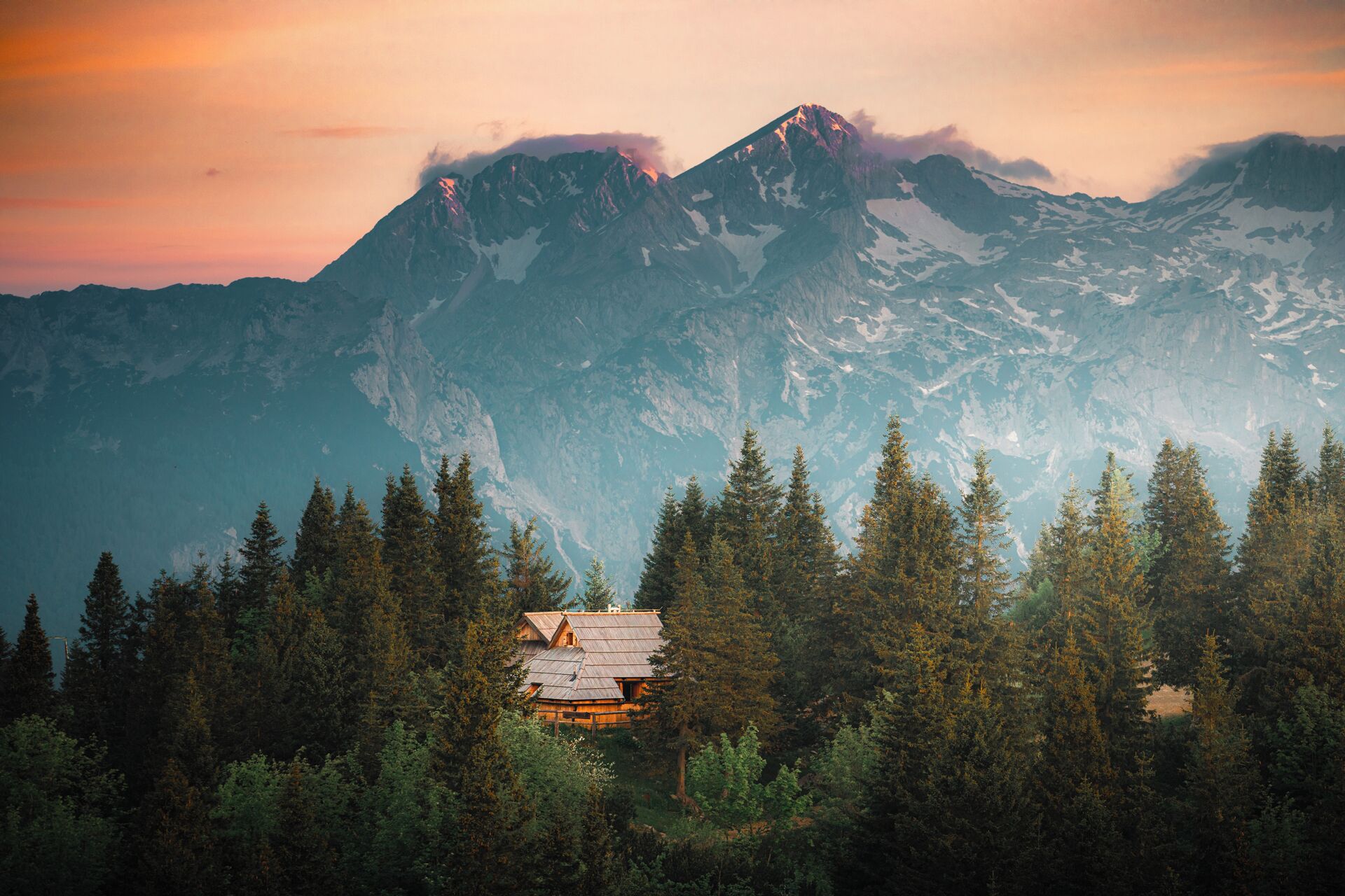 Una cabaña rodeada de un bosque denso con montañas nevadas al fondo.