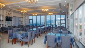 Un comedor amplio con varias mesas y sillas, ventanas grandes que ofrecen vistas a la ciudad y lámparas decorativas.