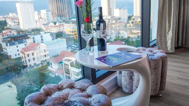Una habitación moderna con una mesa blanca curva, dos copas de vino, una botella y un jarrón con flores. Un otomano mullido y un libro están sobre la mesa. Un ventanal grande ofrece vistas de la ciudad.