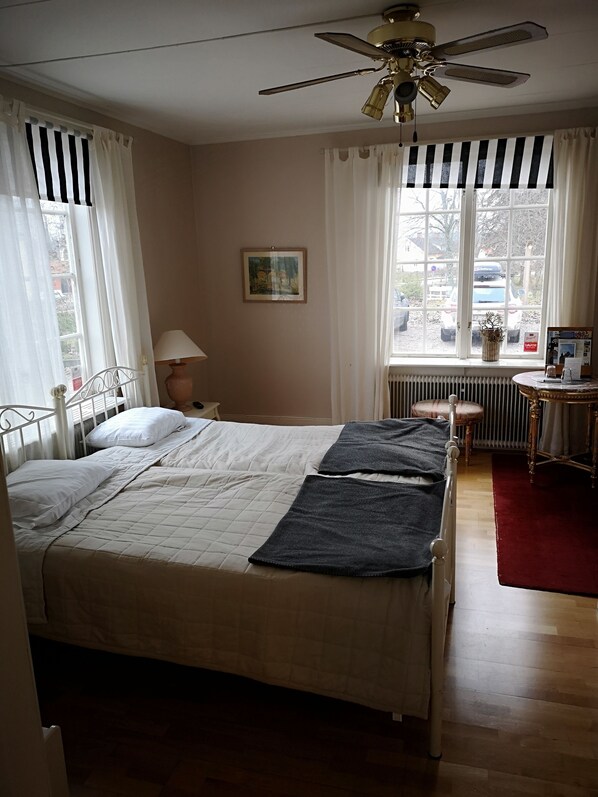 Un dormitorio con cama, ventilador de techo, ventana con cortinas, radiador y escritorio con silla.