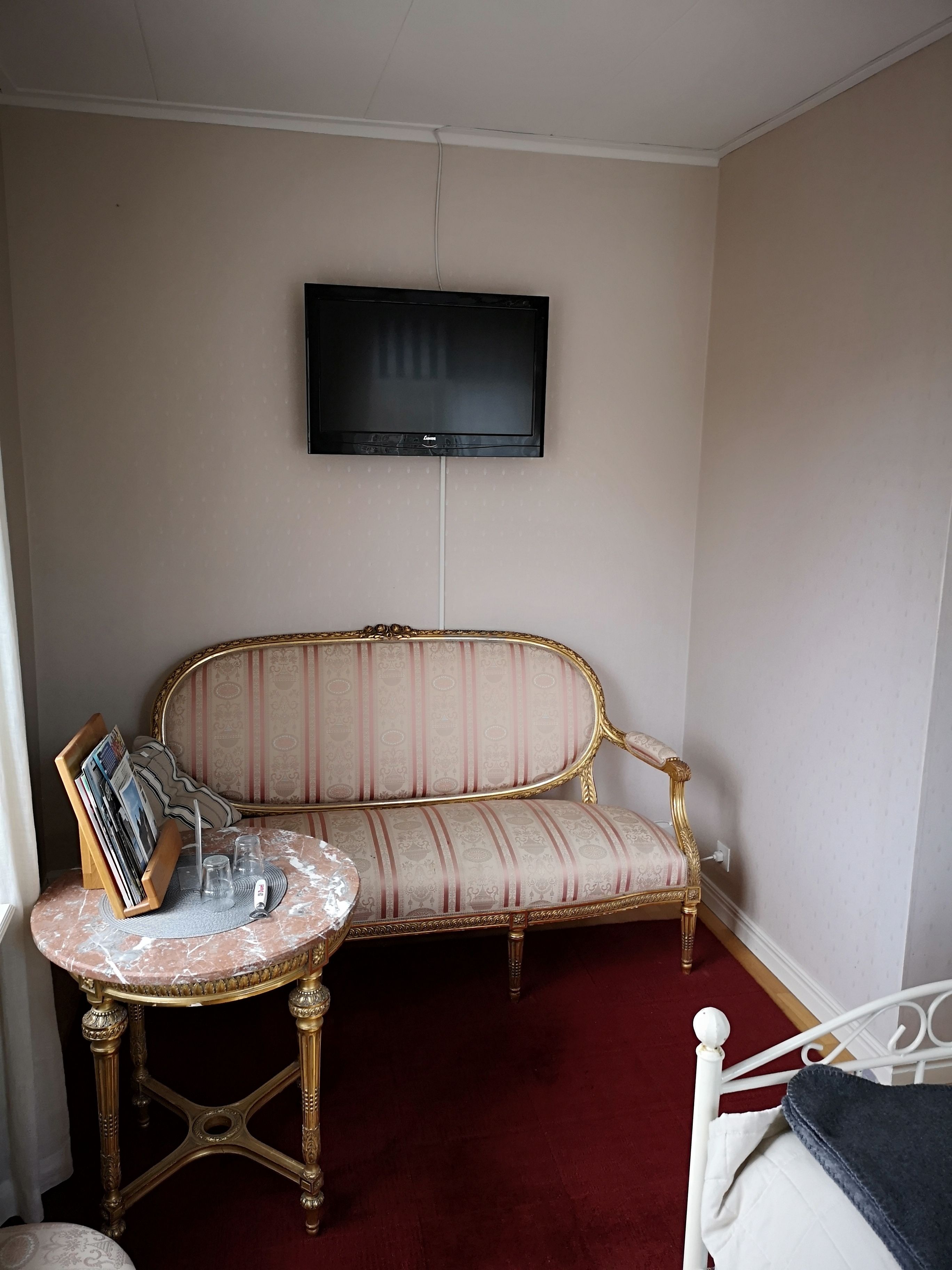 A room with a sofa, a small table with magazines and glasses, a television on the wall, and a bed with a blanket.