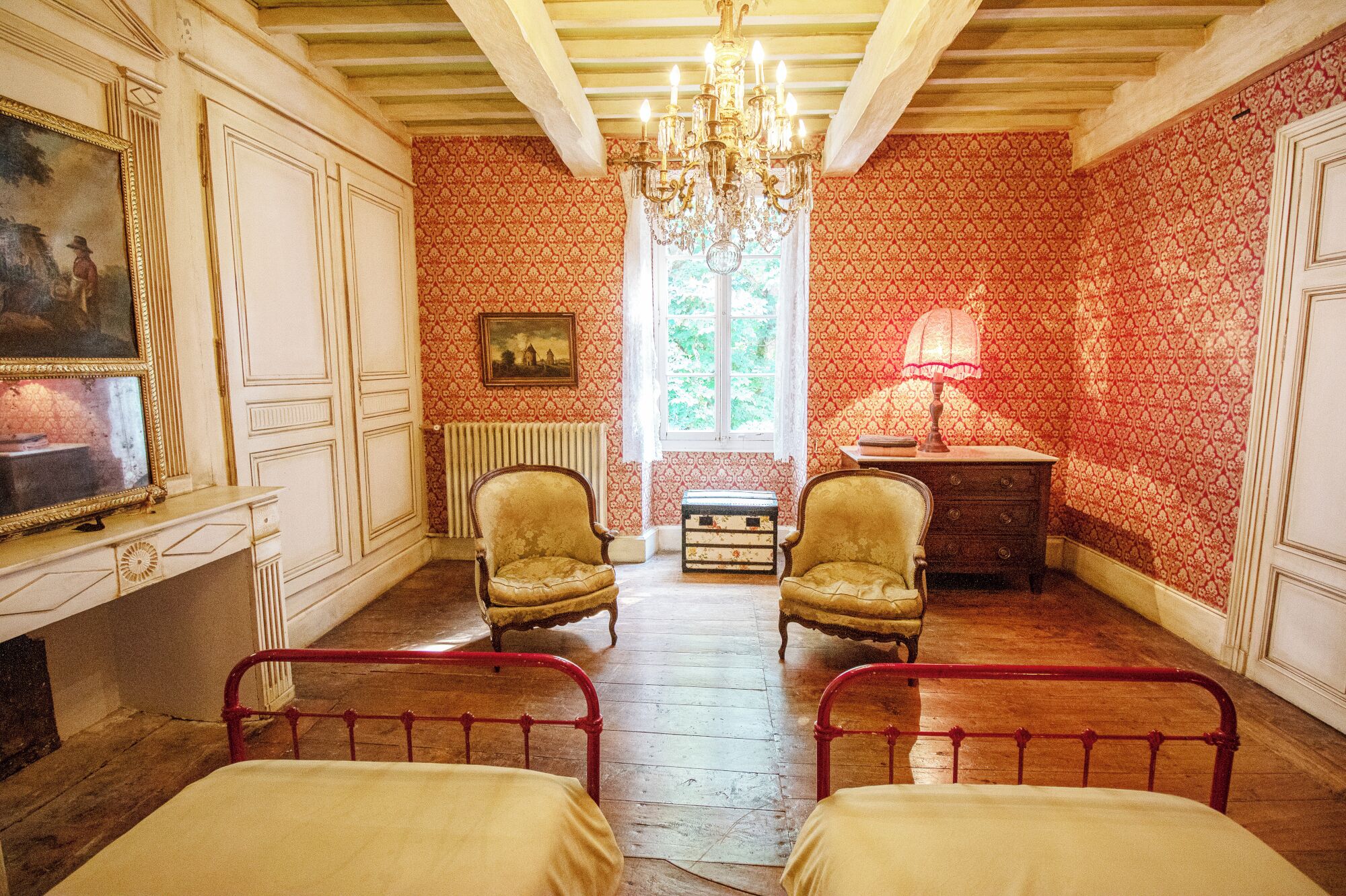 Een kamer met twee bedden, een kroonluchter, een radiator en een raam met uitzicht op groen.