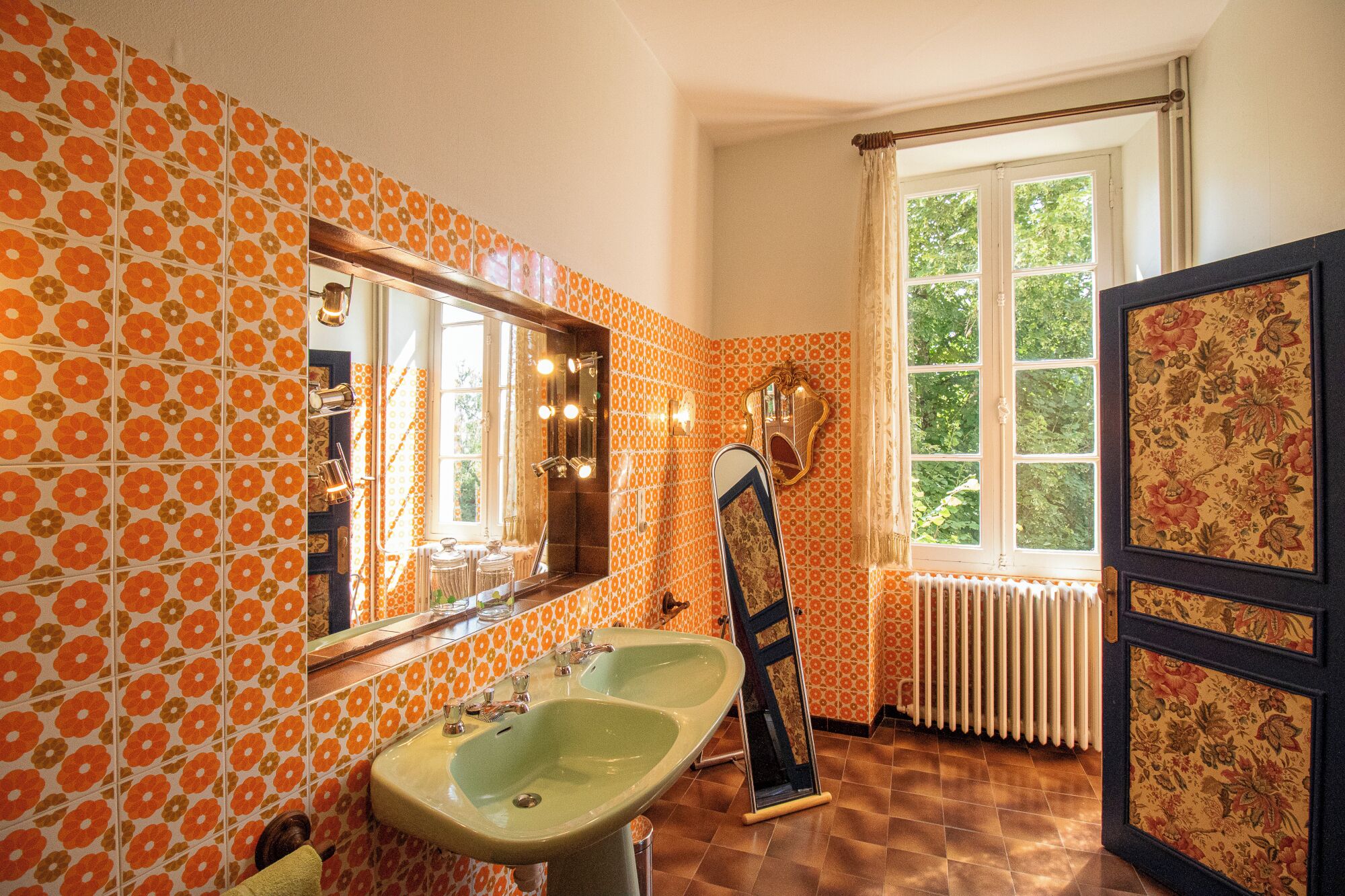Une salle de bain avec des carreaux de céramique à motifs floraux orange, un grand miroir, deux lavabos et une fenêtre donnant sur un paysage verdoyant.