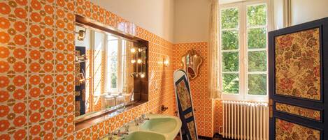 Une salle de bain avec des carreaux de céramique à motifs floraux orange, un grand miroir, deux lavabos et une fenêtre donnant sur un paysage verdoyant.