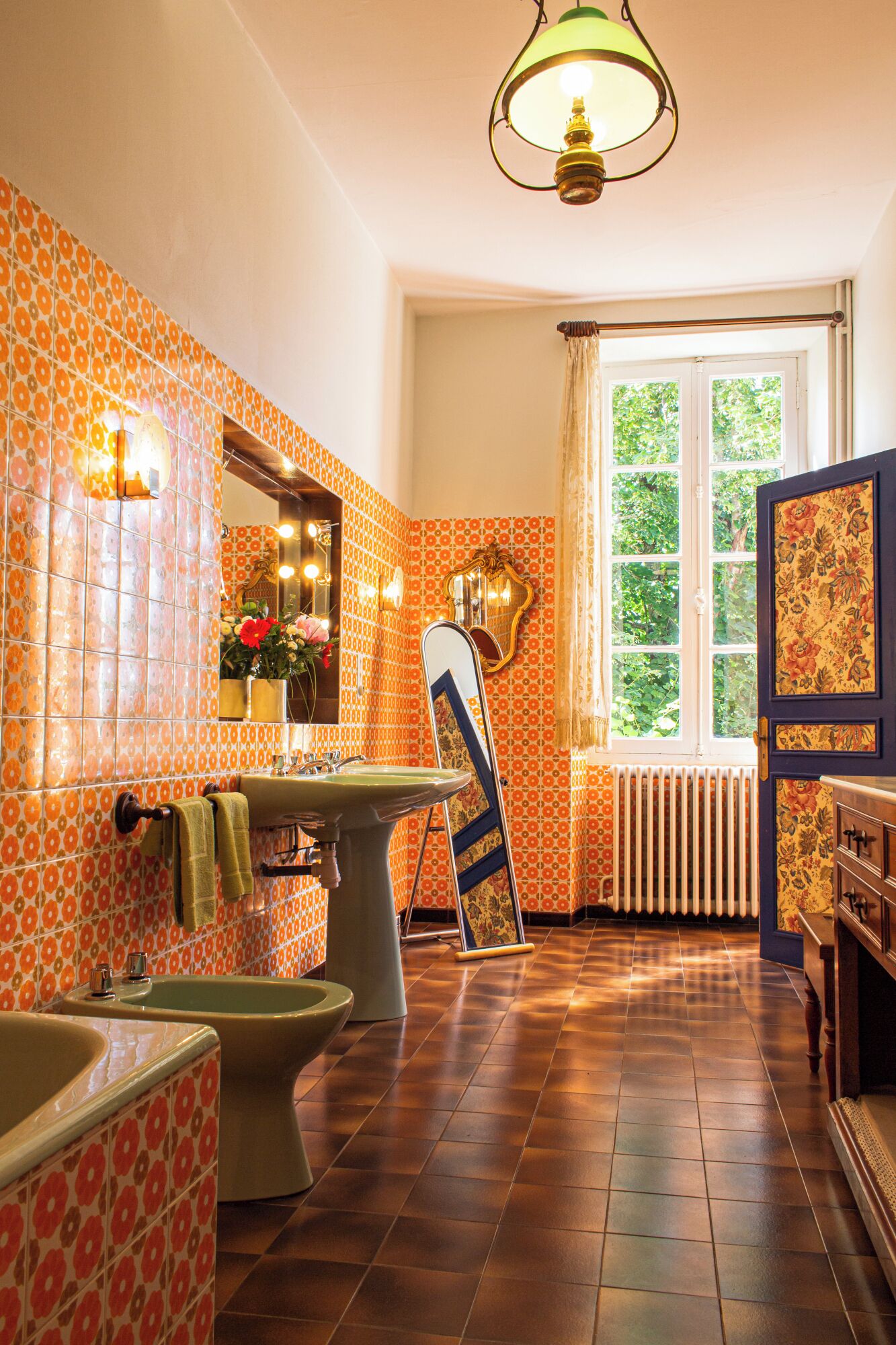 A bathroom with patterned tiles, a sink, a mirror, a radiator, and a window with a view of greenery.