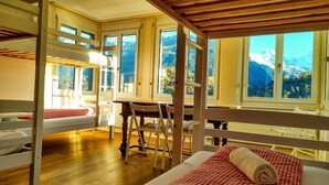 A dormitory-style room with bunk beds, a wooden table, and chairs. Large windows offer views of mountains.