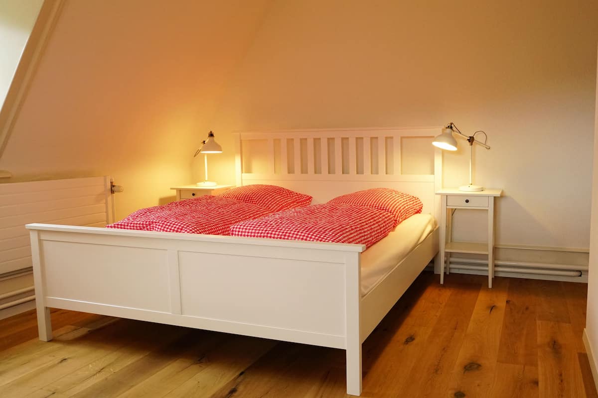 A white bed with red checkered bedding in a room with wooden flooring and white walls.