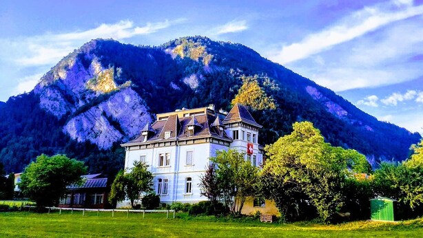 A large, historic building with multiple windows and a prominent tower, surrounded by lush greenery and a mountain backdrop.