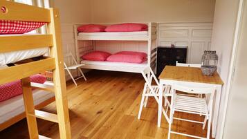 Een slaapkamer met stapelbed, een houten tafel en stoelen, een klein kastje en een raam met luiken.