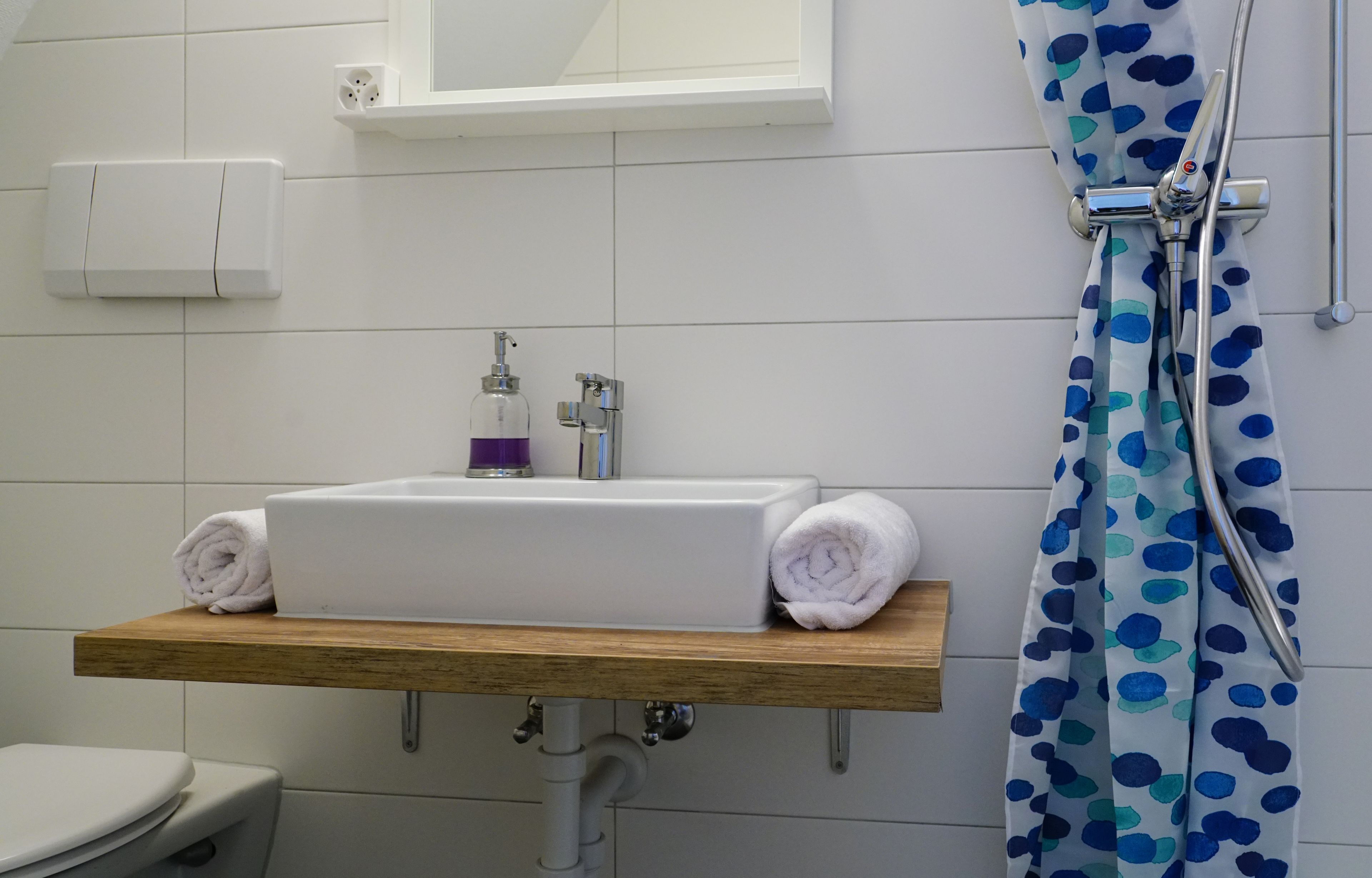 Une salle de bain avec un lavabo blanc, une étagère en bois, une serviette, un rideau de douche et un miroir.