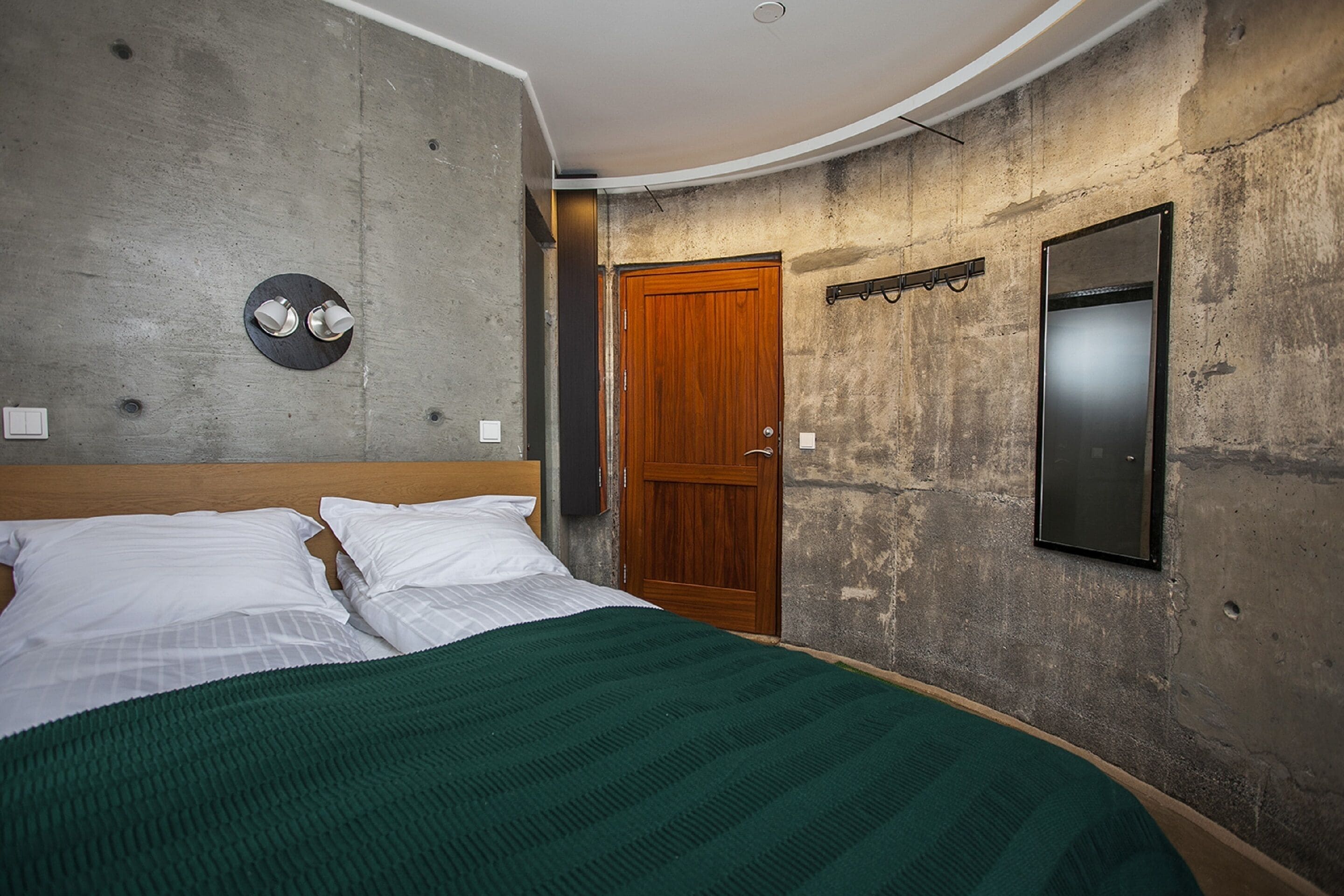 A bedroom with a bed, a wooden door, a wall-mounted mirror, and a circular light fixture.