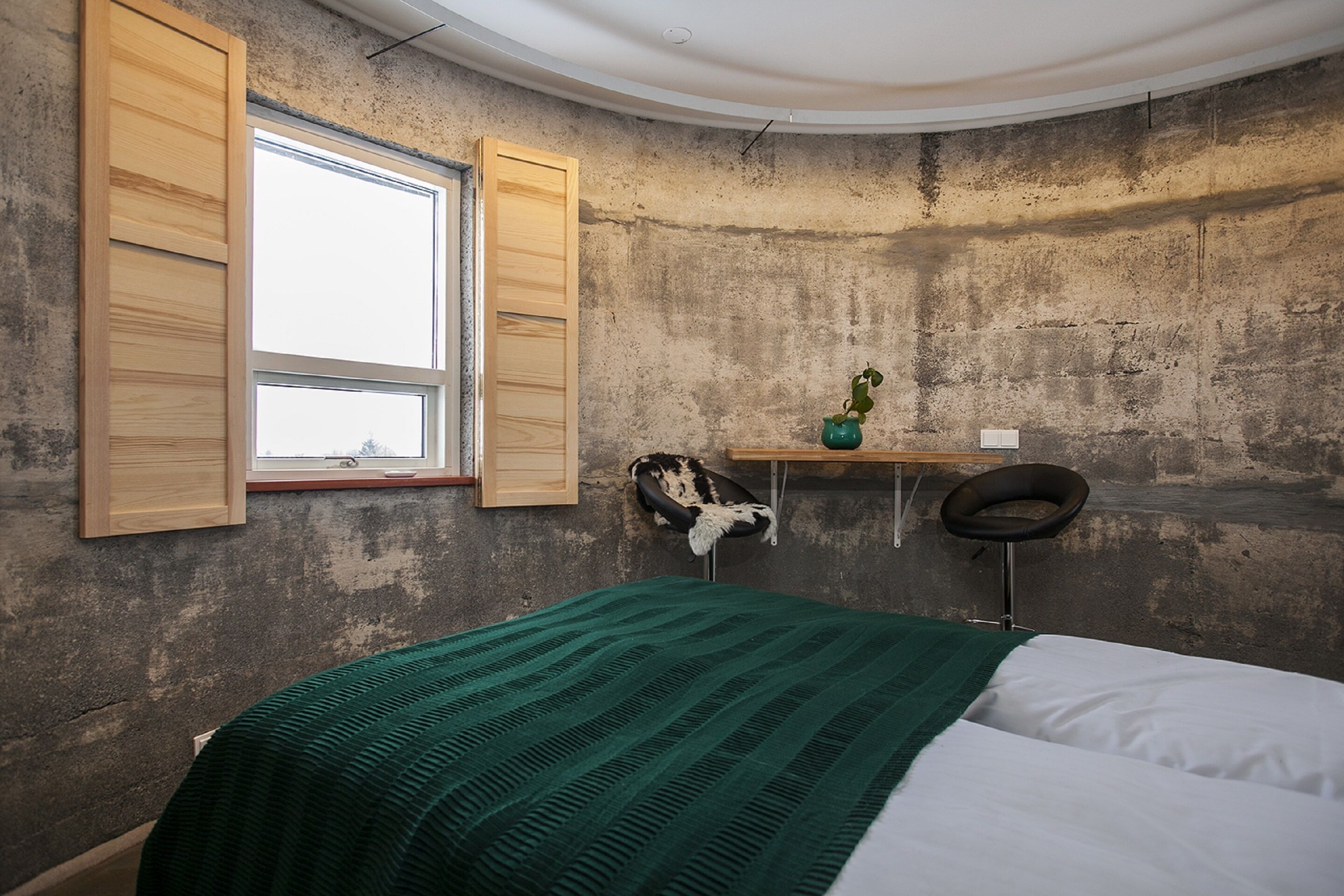 A bedroom with a bed, a window with wooden shutters, a desk with a chair, a potted plant, and a wall-mounted shelf.
