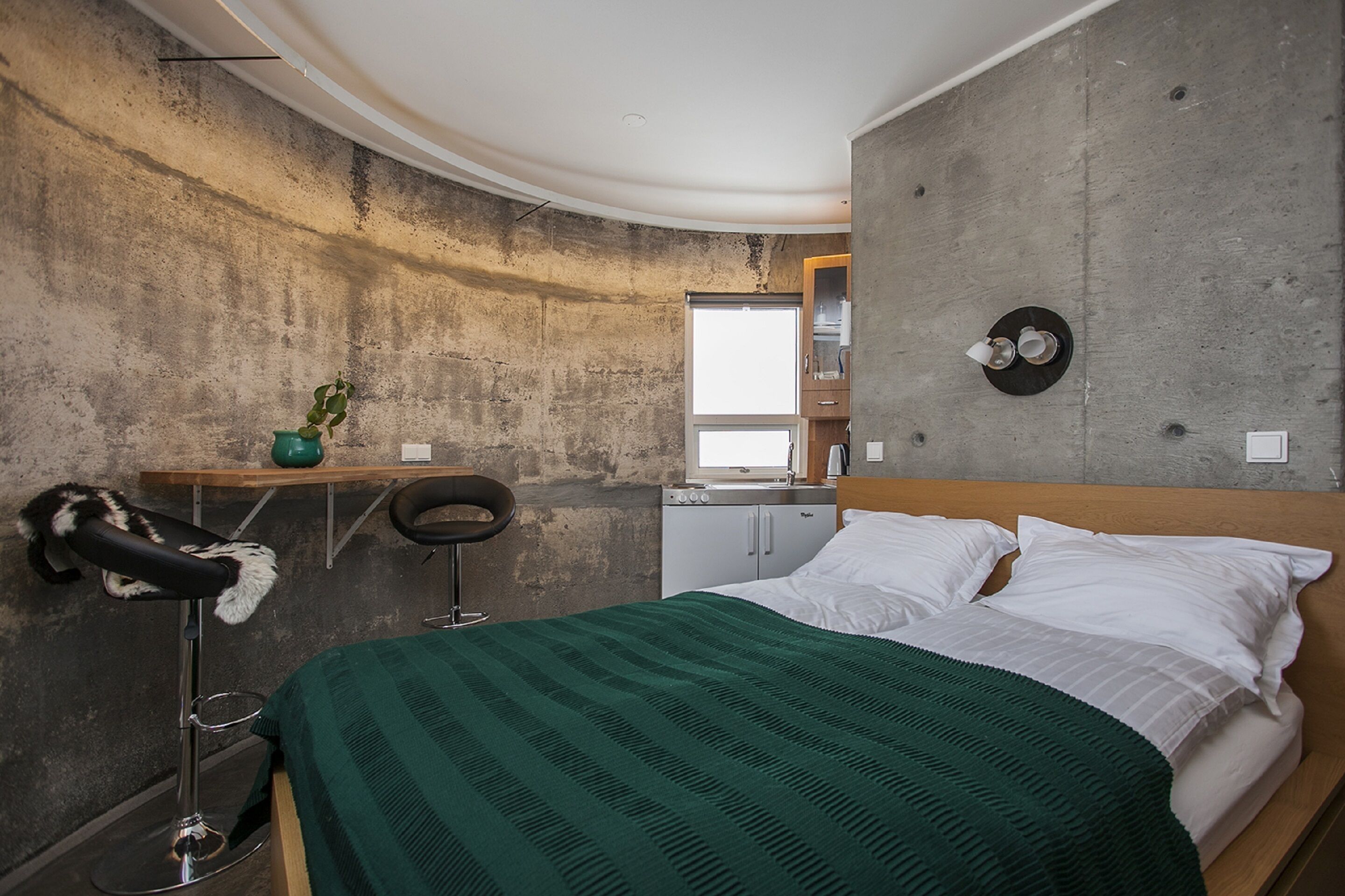 A compact bedroom with a bed, a small desk, a chair, a potted plant, and a window.