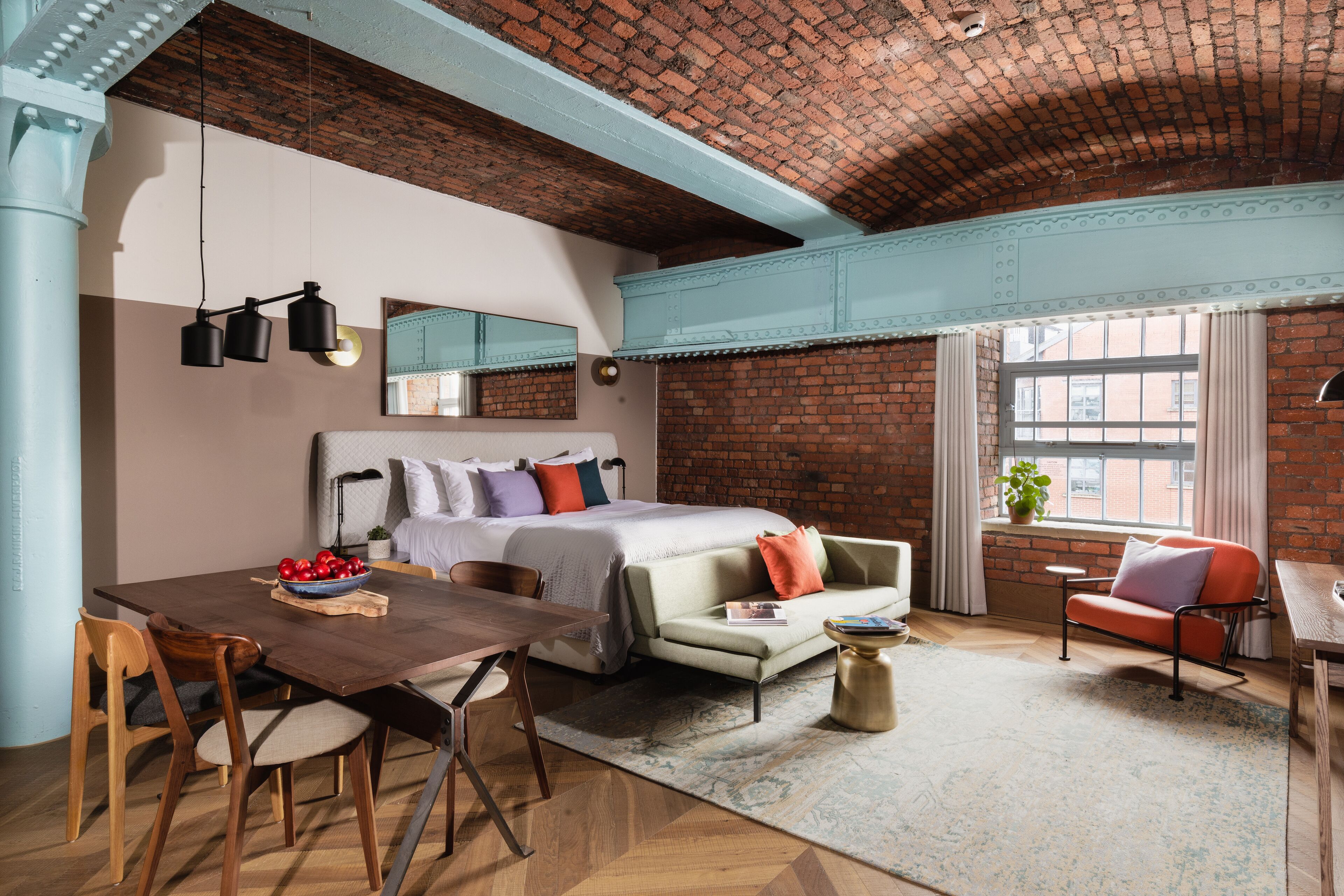 A bedroom with a brick wall, a bed, a nightstand, a window with curtains, and a dining area with a table and chairs.