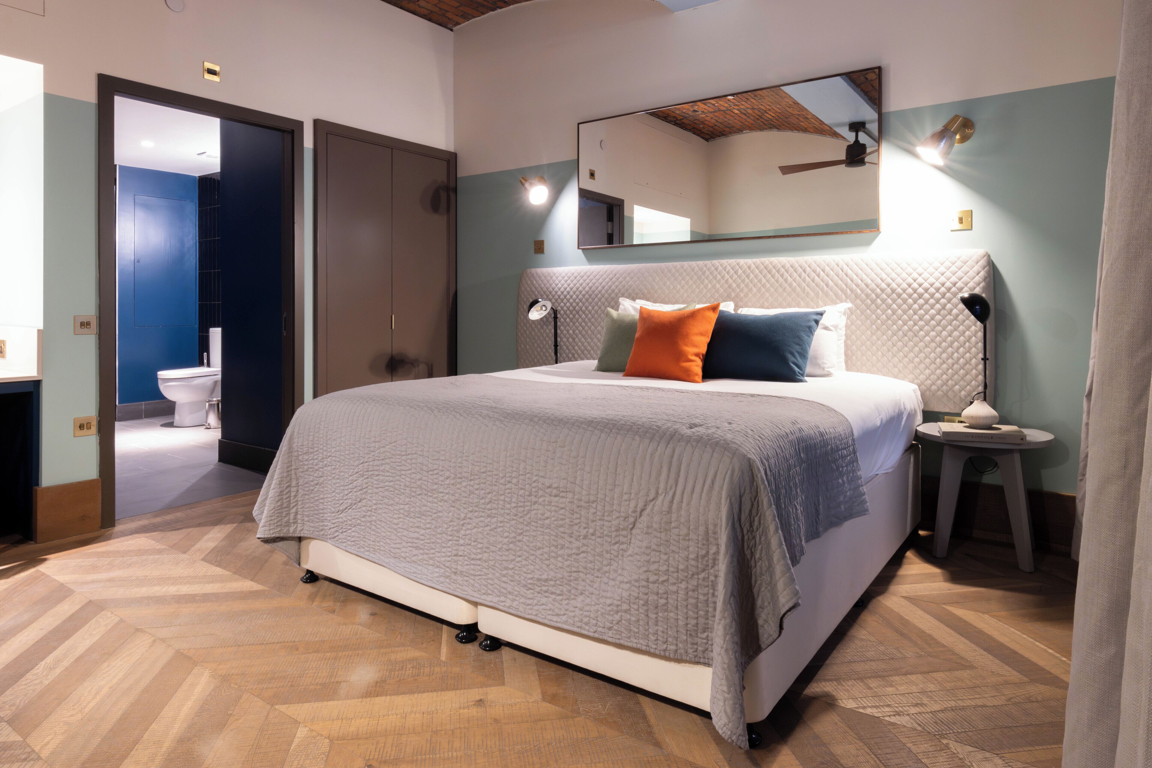 A modern bedroom with a large bed, a bedside table, a nightstand, a mirror, and a door leading to a bathroom.