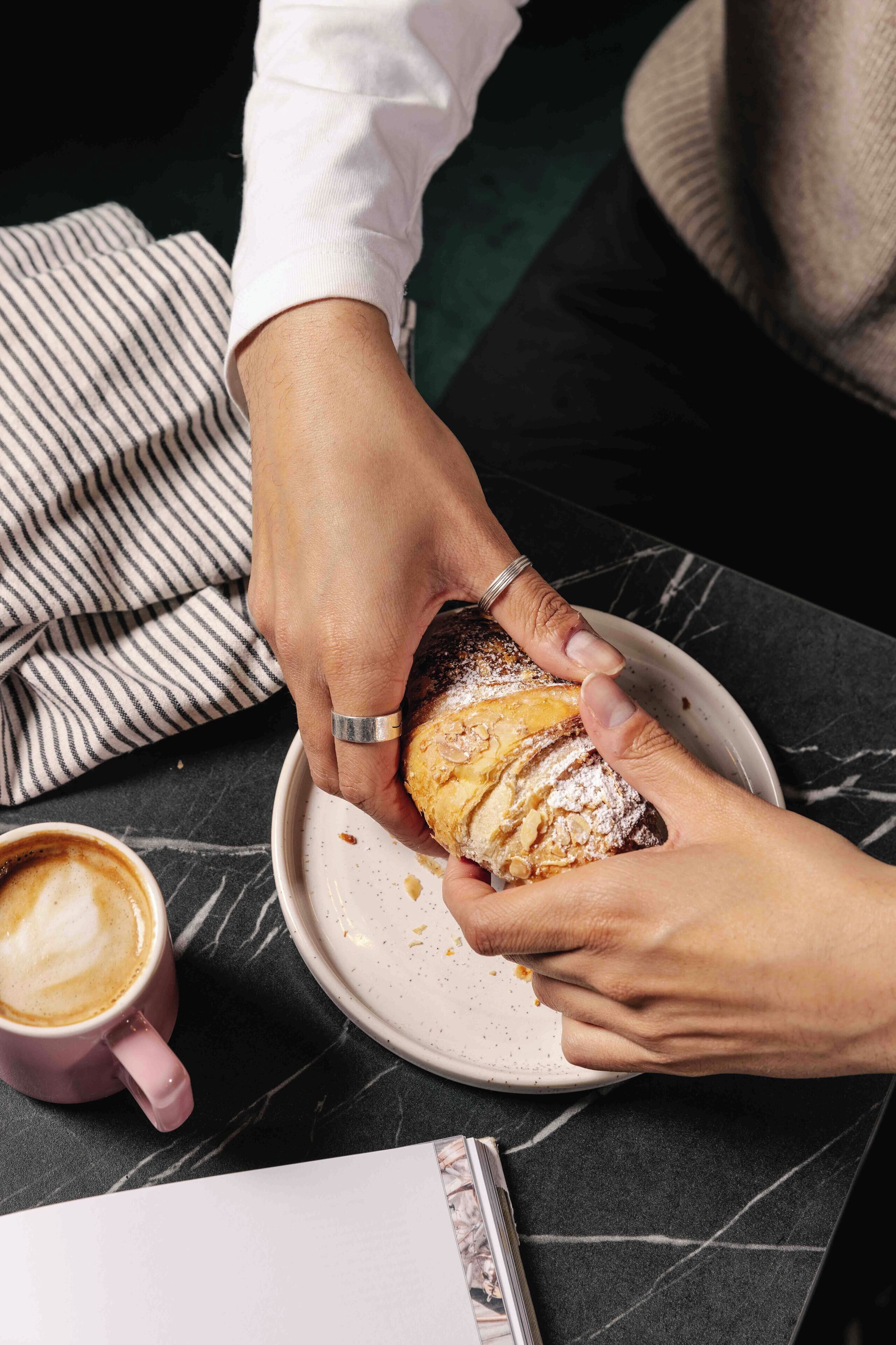 Une personne tenant une pâtisserie saupoudrée de sucre glace sur une assiette, une tasse de café, et un tissu à rayures en arrière-plan.