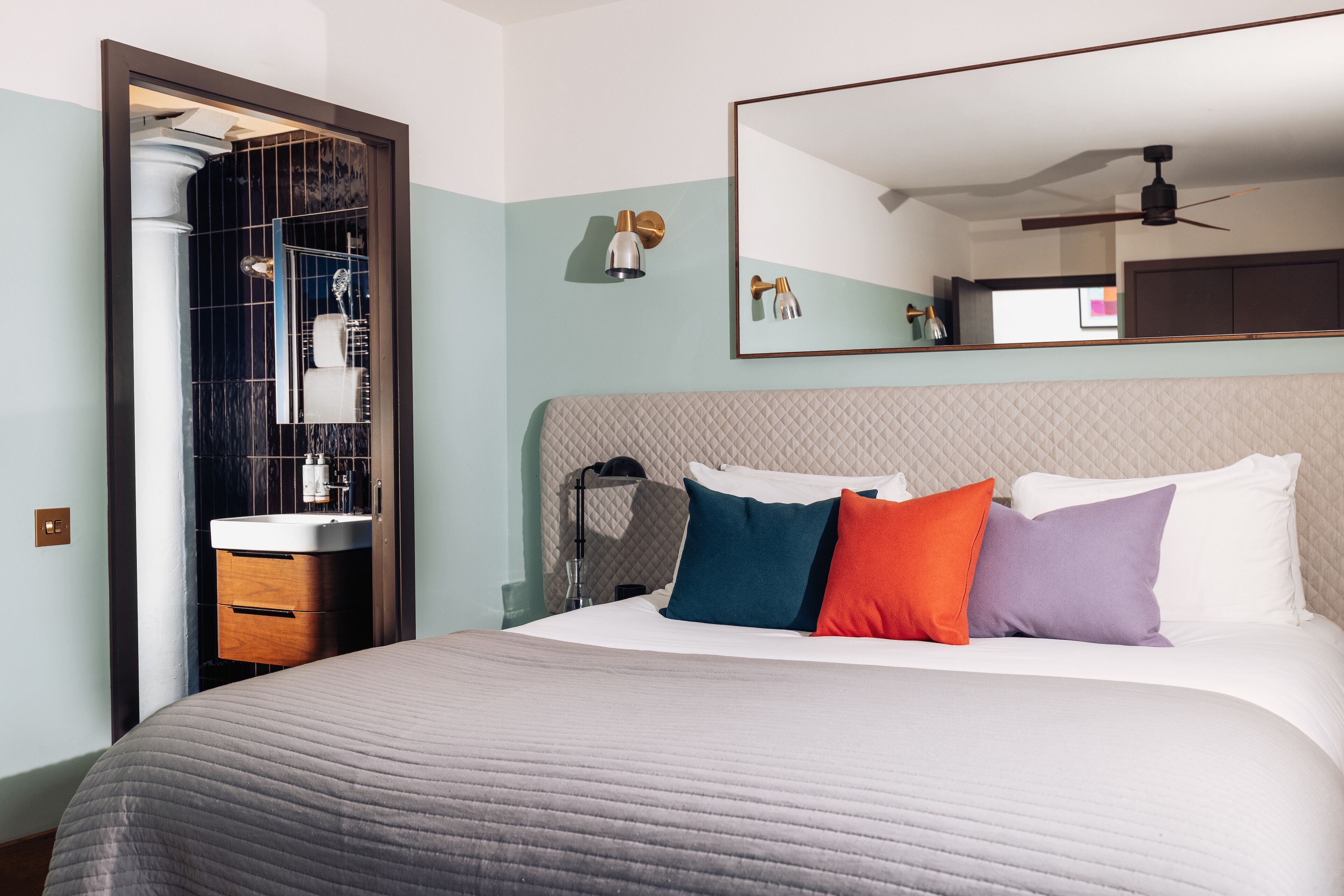 Une chambre d’hôtel comprenant un lit, une table de chevet, un miroir, un ventilateur de plafond et une salle de bain avec un lavabo et un miroir.