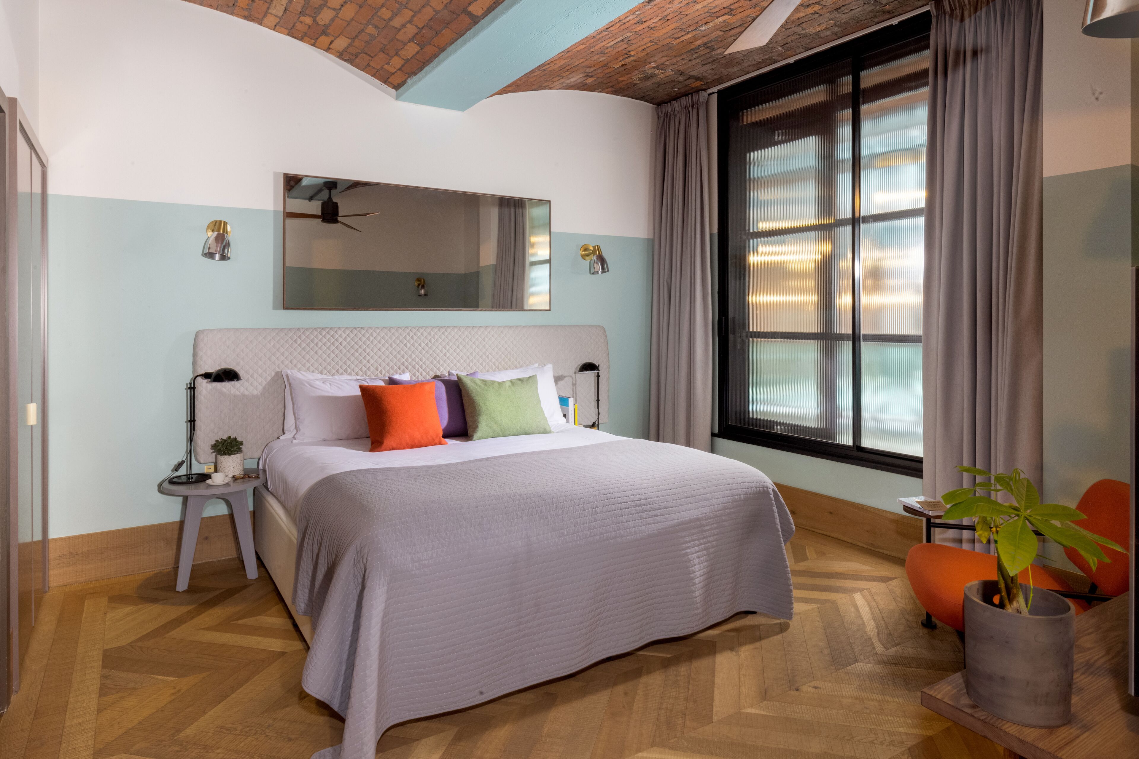 Quarto moderno com uma cama grande, um espelho acima, uma janela com cortinas, uma mesinha com uma planta e uma cadeira.
