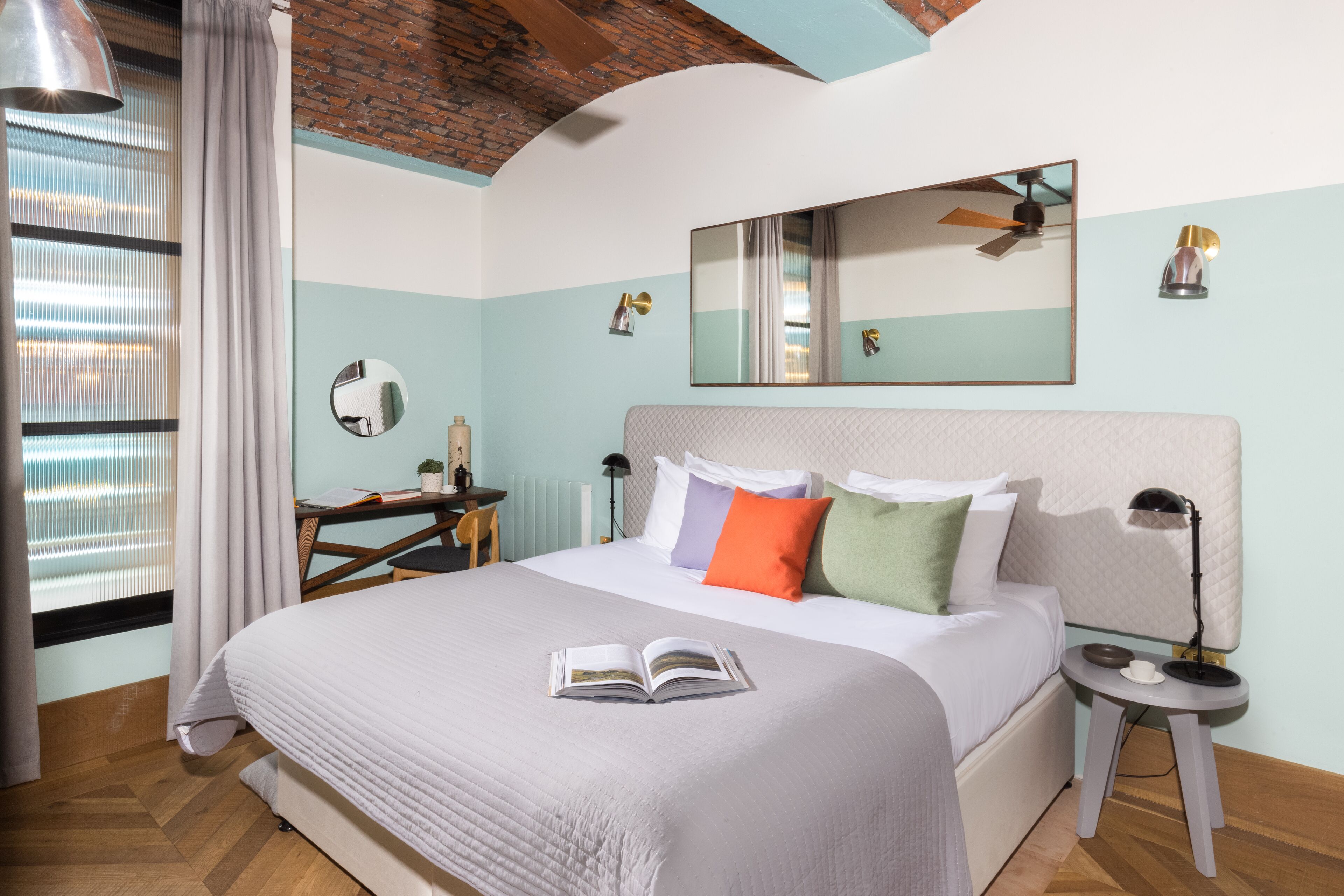 A neatly made bed with a book, a desk with a chair, a mirror, and a bedside table with a lamp.