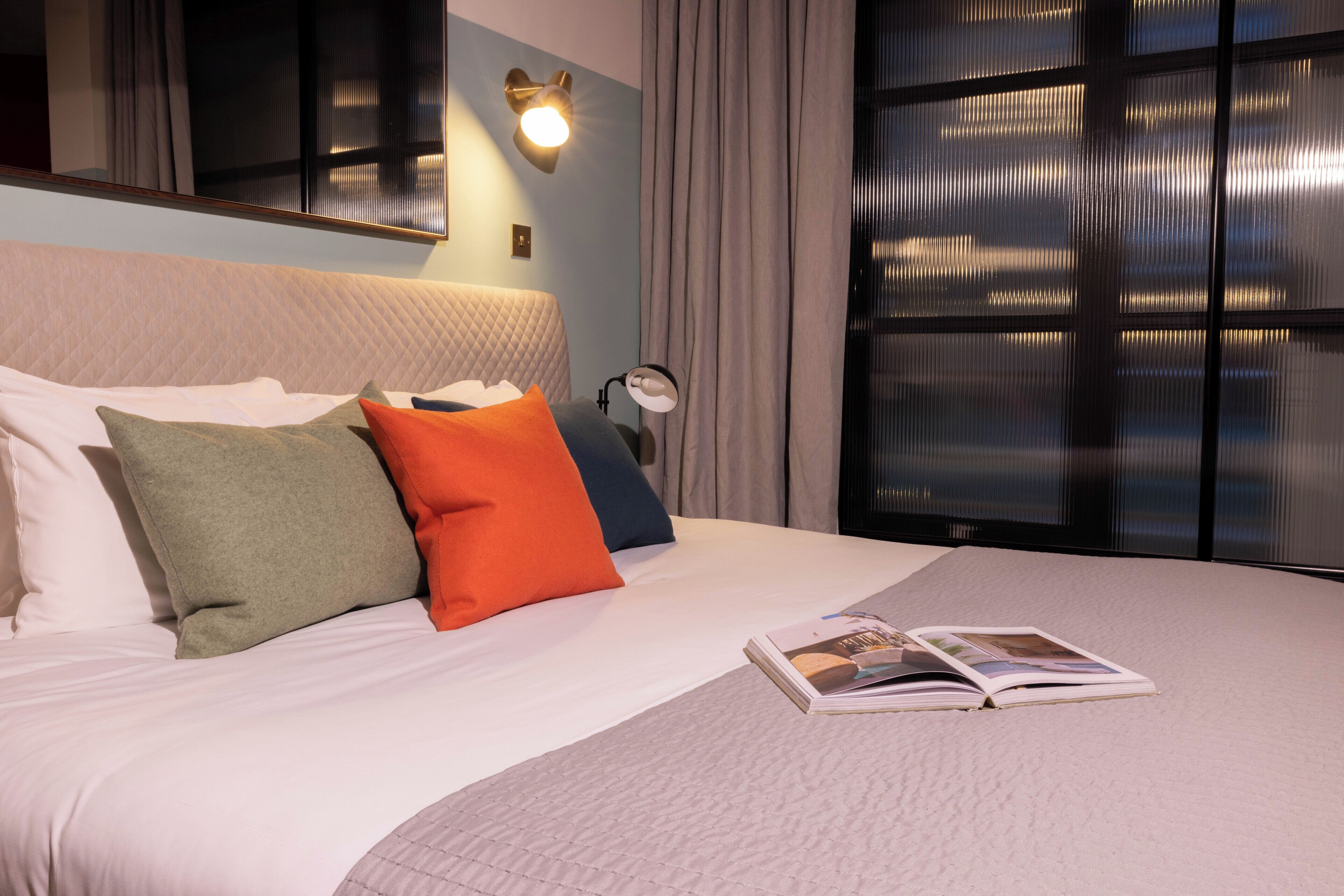 A neatly made bed with a magazine, pillows in various colors, a wall-mounted lamp, and a large window with curtains.