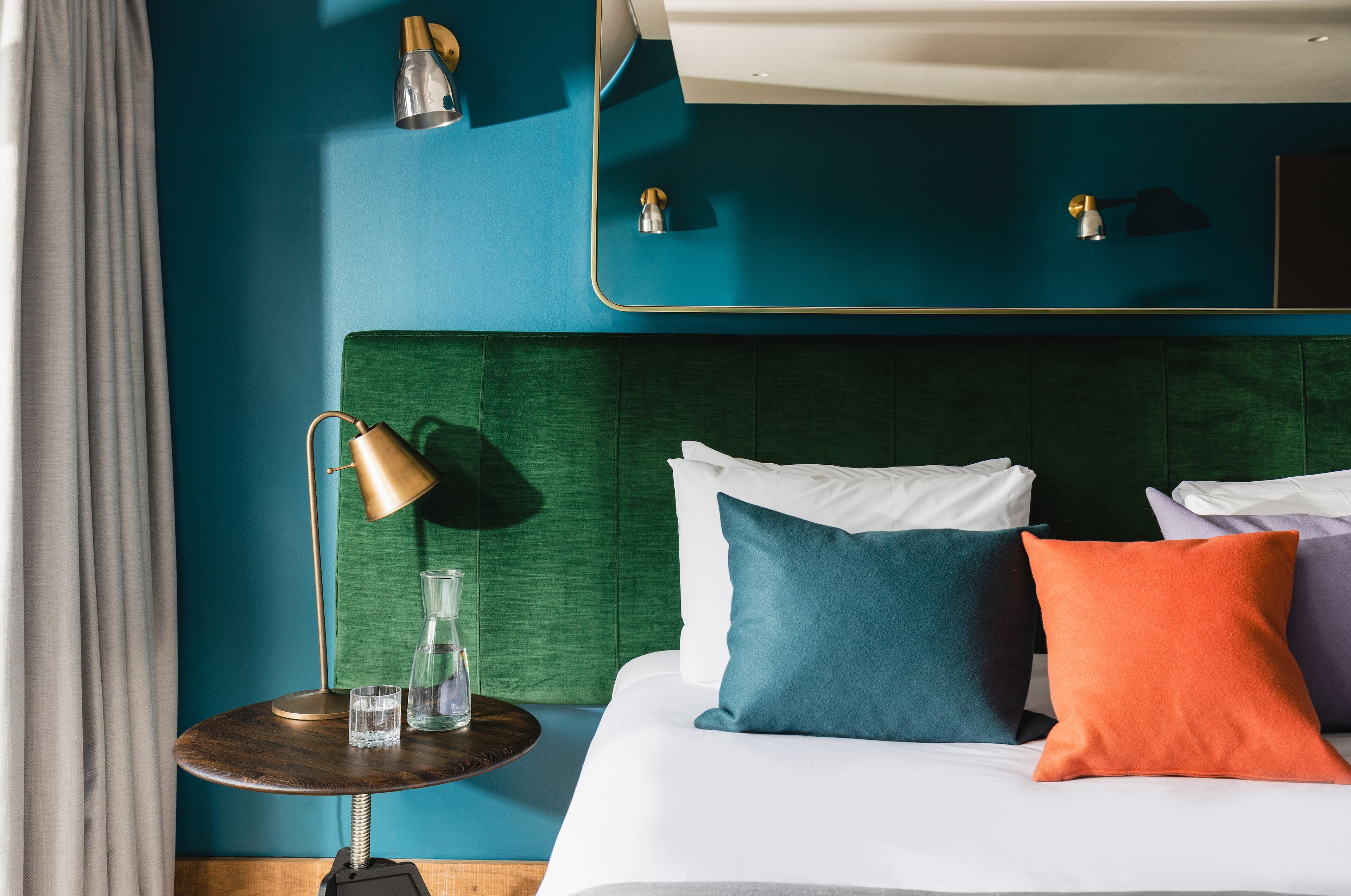 Une chambre d’hôtel avec une tête de lit recouverte d’un tissu vert, du linge de lit blanc et une petite table ronde agrémentée d’une lampe et d’un vase en verre.