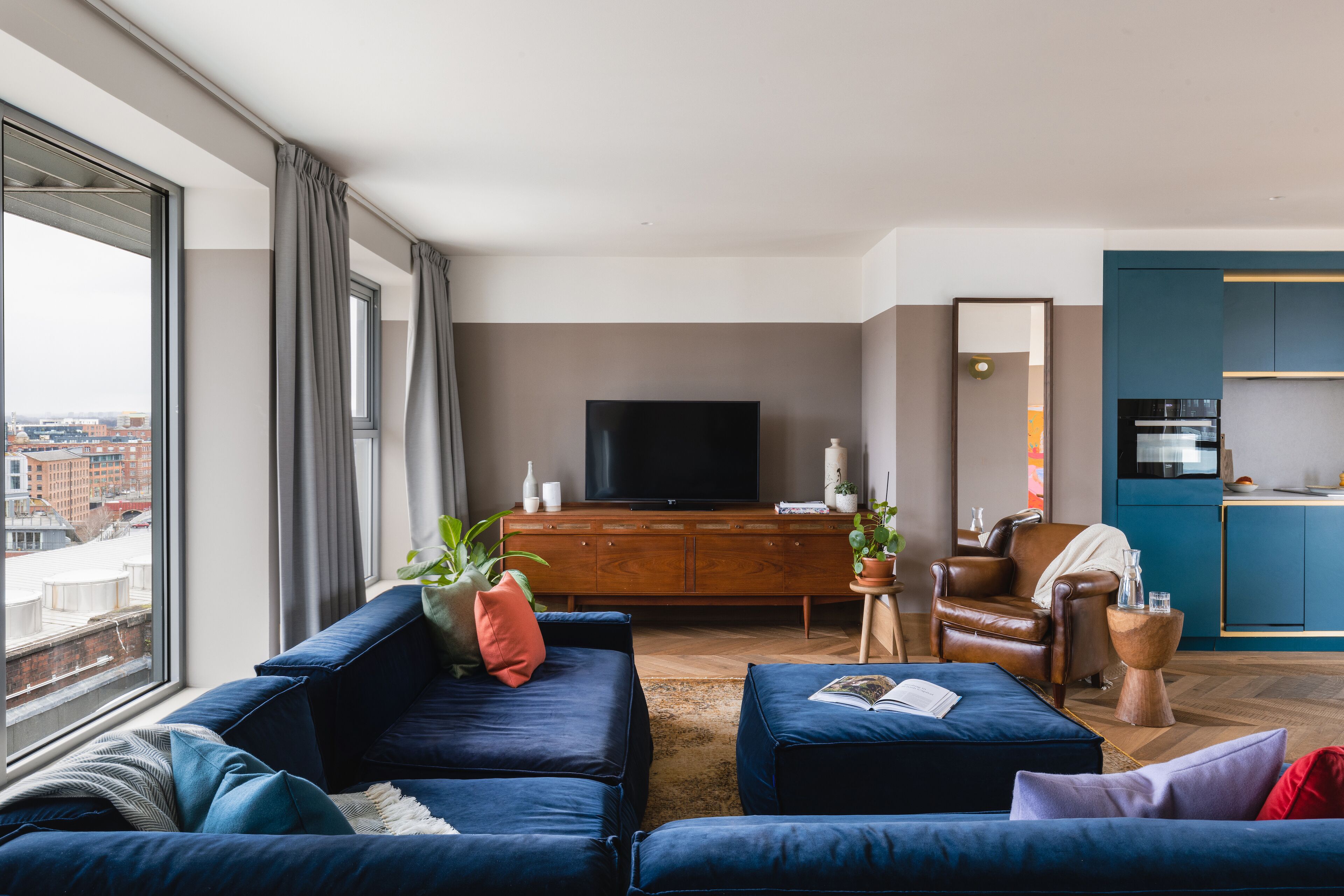 A modern living room with a large window, a flat-screen TV, a brown leather armchair, and a blue sofa set.